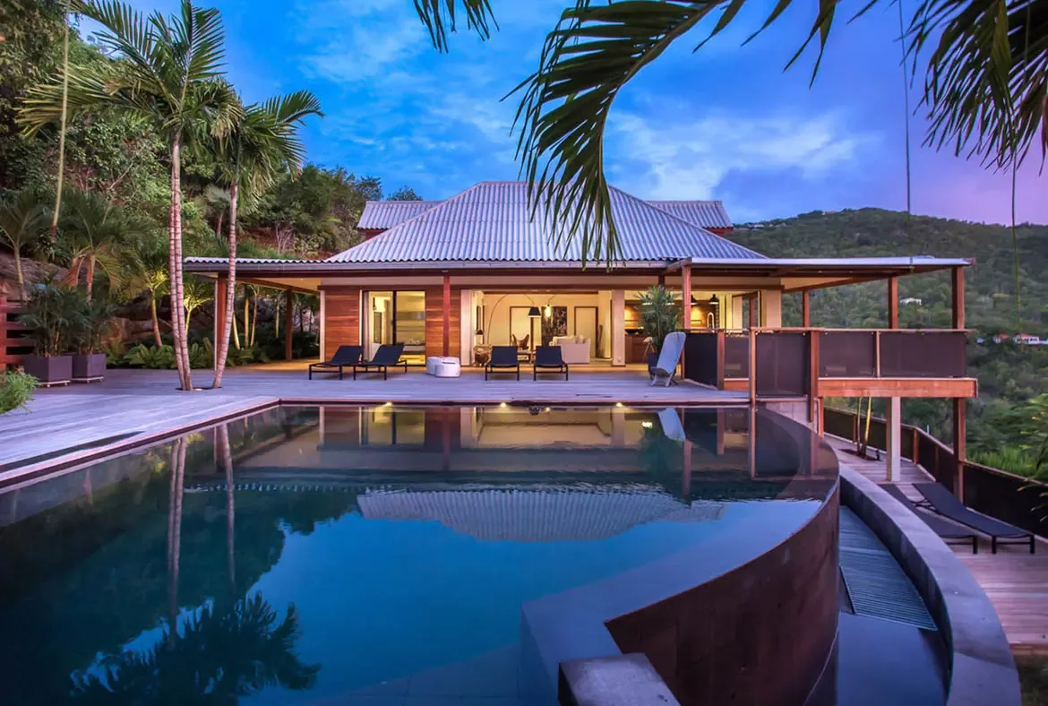 An infinity pool with turquoise water sits beside white loungers on a spacious terrace, where a modern white villa with open-concept living opens to lush tropical hills beneath puffy clouds.