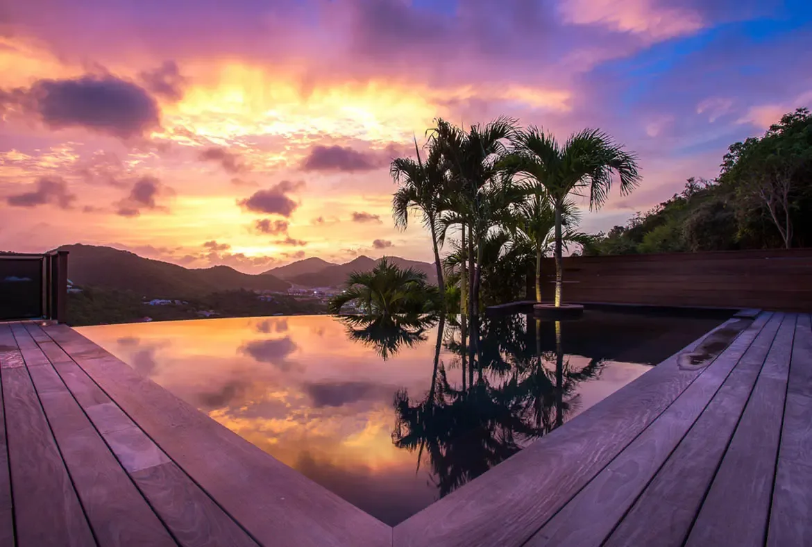 A blue-lit infinity pool reflects a white villa at night, where palm trees and warm interior lights create an elegant evening atmosphere against a dark hillside.