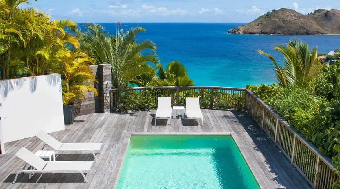 A dark wood bed with white linens sits before open glass doors, where a private terrace leads to a turquoise ocean view beneath wispy clouds.