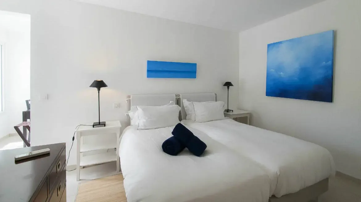 A white bed with plush pillows sits beside open glass doors, where a private infinity pool and lush tropical gardens create a serene bedroom retreat.