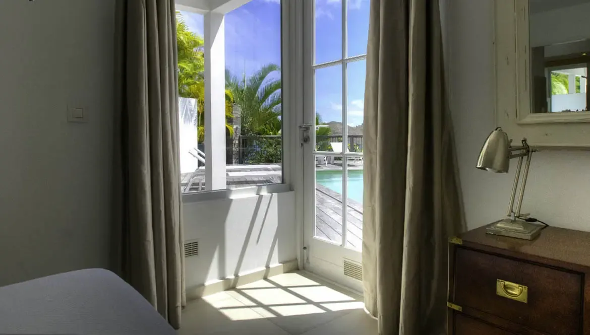 An infinity pool with white loungers sits on a white terrace, where a modern villa with open glass doors overlooks a lush green hillside beneath a bright blue sky.