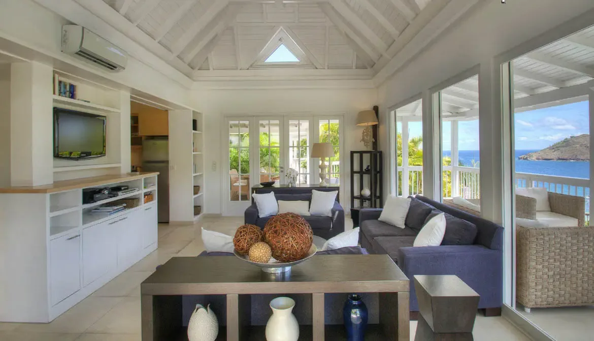 A white dining table with modern chairs sits on a covered terrace, where white pillars and lush tropical gardens create an elegant outdoor dining space.
