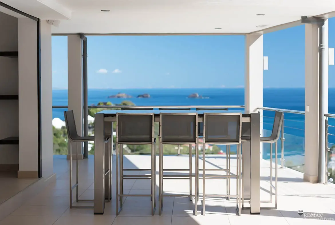 A sleek bar-height dining setup takes full advantage of the terrace, with uninterrupted views of turquoise water and scattered islands beyond. Bright sun, clean lines, and a glass railing keep the focus exactly where it belongs, on the Caribbean horizon.
