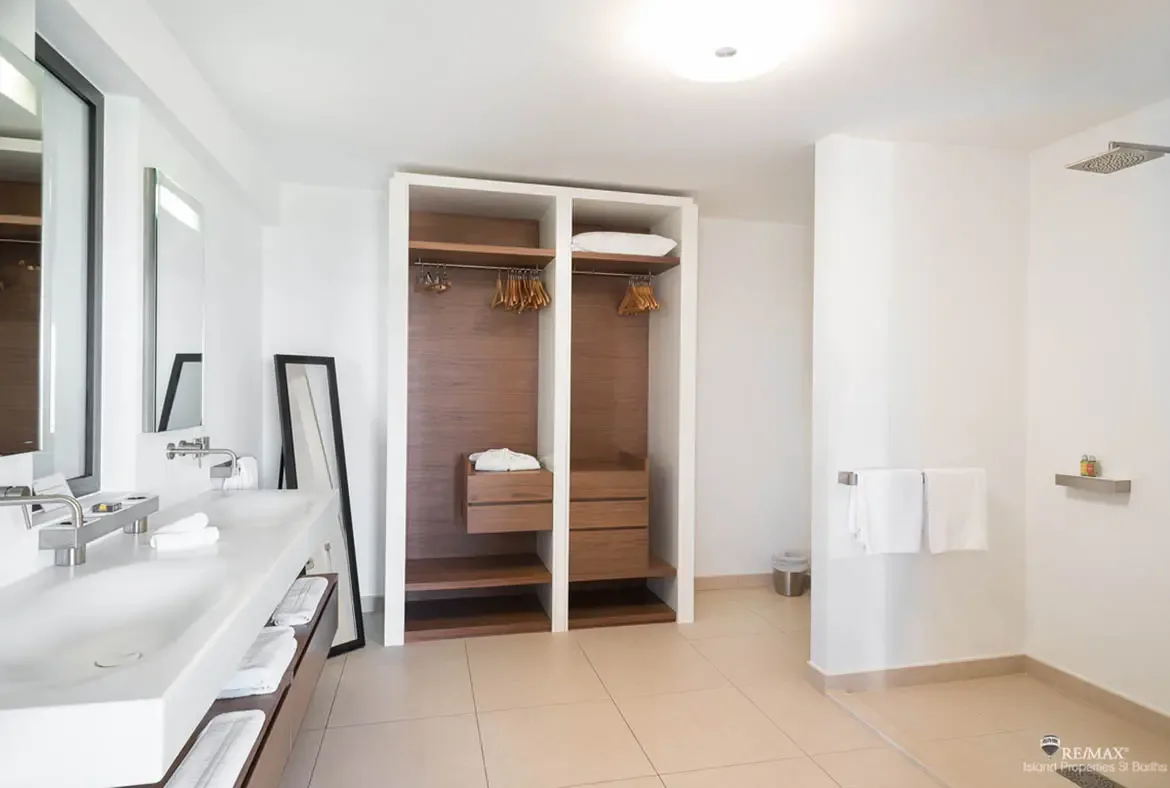 A long double-sink vanity flows into an open dressing area with warm wood shelving and neatly lined hangers, giving this suite a polished, boutique feel. Crisp white finishes and clean geometry keep the space calm, bright, and easy to settle into.