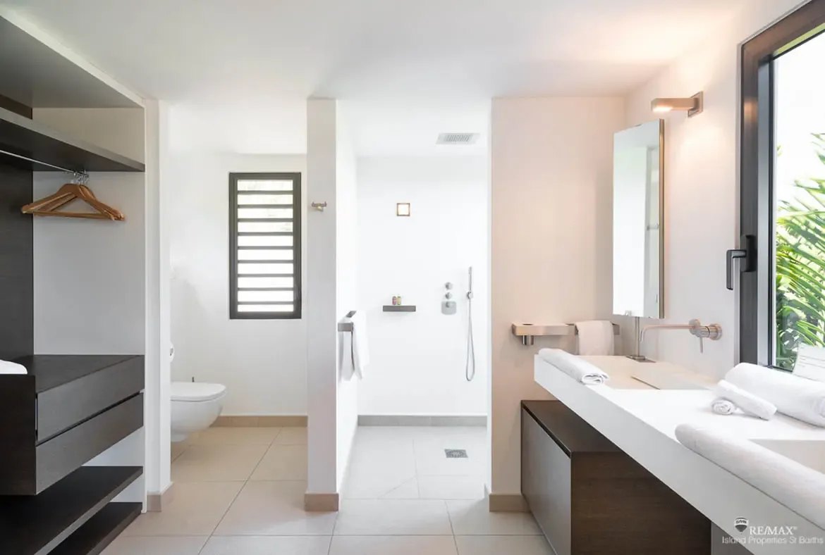 This bright open-plan bathroom pairs a sleek white vanity and walk-in shower with a dark wood dressing area for a polished, contemporary feel. A large window pulls in lush tropical greenery, softening the clean lines with a sense of island calm.