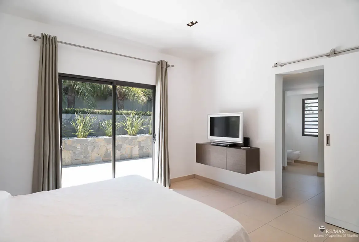 This bright bedroom keeps the mood calm and minimal, with white bedding, soft gray curtains, and a glass door opening to tropical greenery just outside. The open ensuite beyond adds a clean modern flow that makes the suite feel airy and effortless.