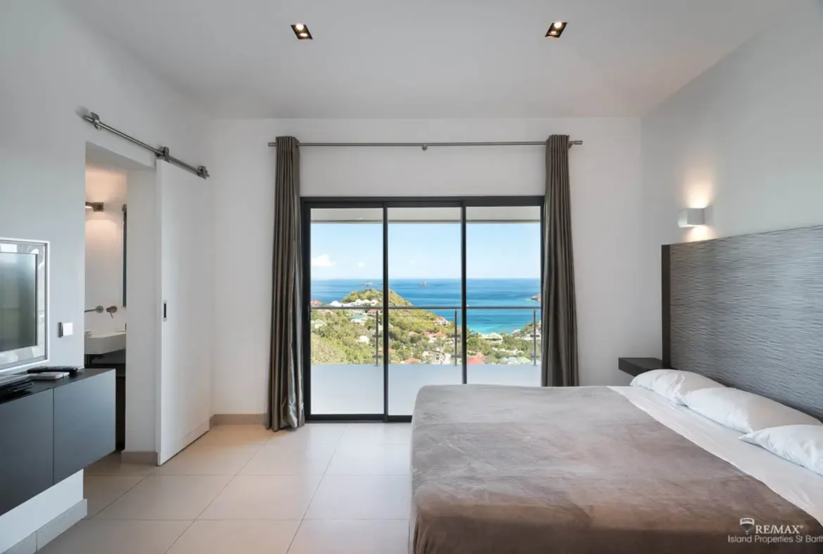 Dark-framed doors open this calm, modern bedroom to a sweeping hillside and ocean panorama, with red-roofed villas scattered below. Soft neutral bedding, clean lines, and a quiet palette let the Caribbean view do exactly what it should, steal the whole scene.