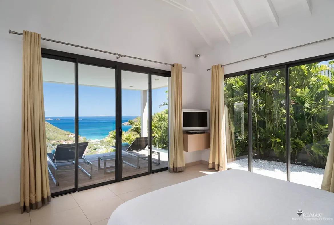 This serene white bedroom opens in two directions, one side framing the turquoise bay and the other a lush private garden. With vaulted beams, soft neutral drapes, and a terrace just beyond the bed, it feels completely immersed in the island landscape.