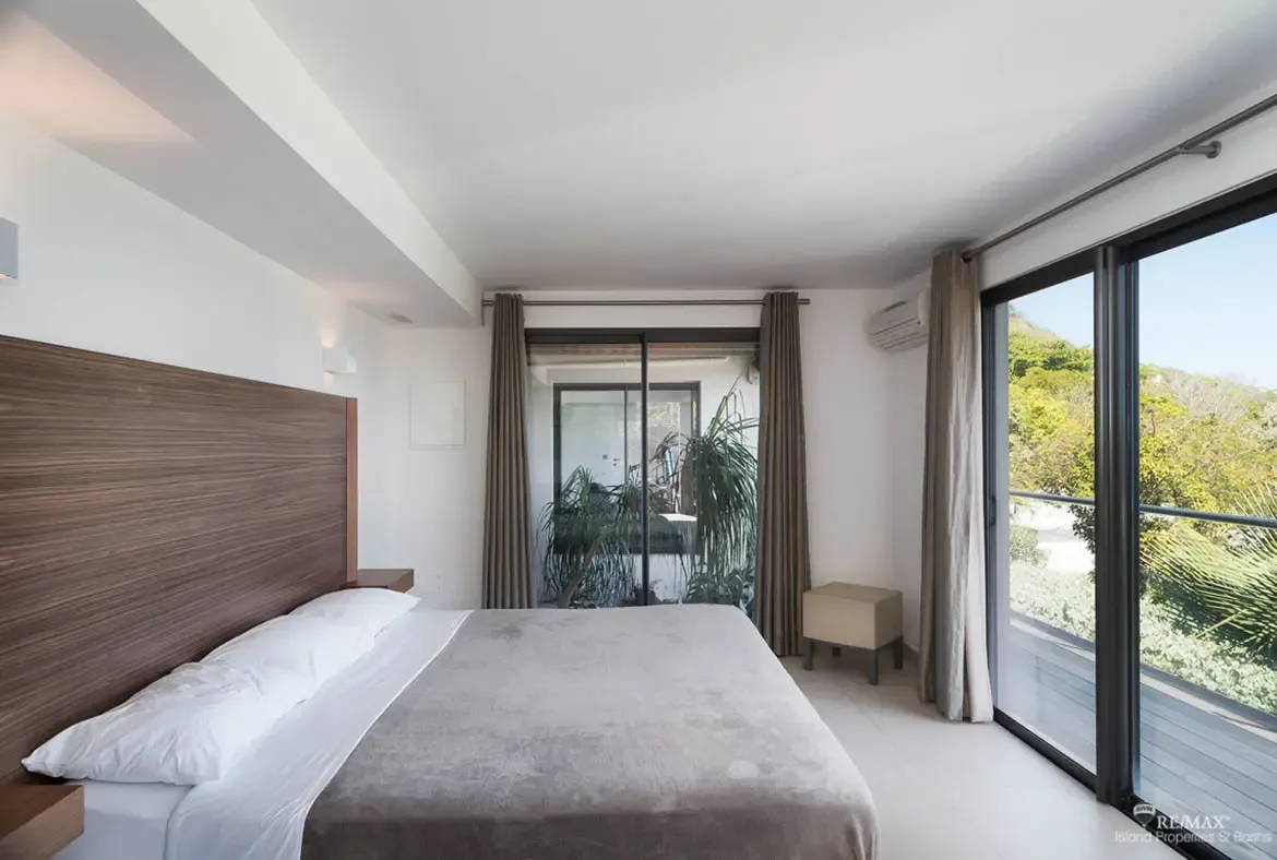 This serene bedroom is wrapped in light from two sides, one opening to a lush tropical atrium and the other to a sunlit hillside terrace. Soft neutral tones, a wide wood headboard, and clean modern lines make it feel calm, private, and effortlessly luxurious.