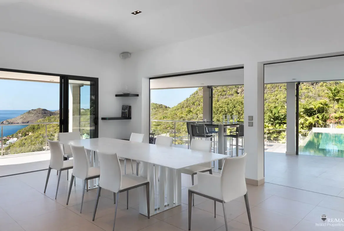 A long white dining table and a sleek breakfast bar with stools make this bright interior ideal for gathering from morning through evening. Glass doors on both sides pull in hillside, pool, and ocean views, keeping the whole space connected to the landscape.