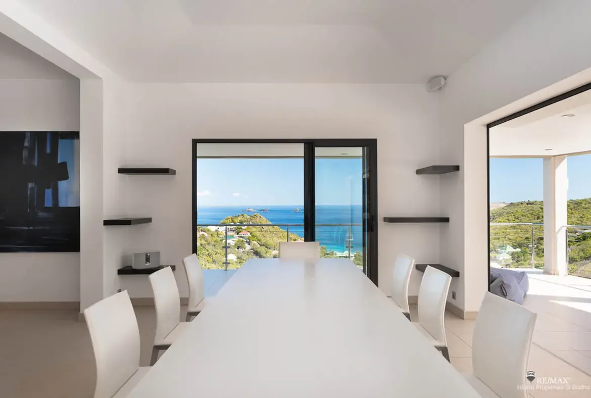 A sleek white dining table for six keeps the mood crisp and contemporary, while floating shelves and abstract art add quiet character. With ocean and green hillside views framed by the windows, even a simple lunch here feels elevated.