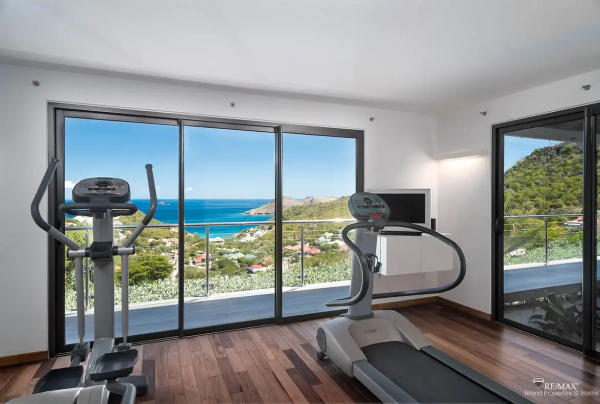 This bright home gym turns every workout into a view-driven escape, with cardio machines facing the hillside and the Caribbean beyond. Glass doors open wide to the terrace, so the room feels less like a gym and more like a private wellness pavilion.