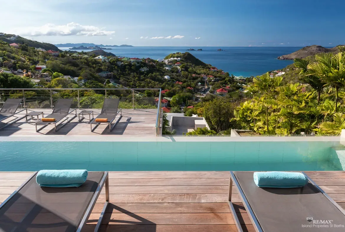 Two loungers with pale blue towels face a long turquoise infinity pool and a panorama of lush hills rolling down to the Caribbean. With villas scattered across the hillside and islands on the horizon, this is one of those views that instantly sells the stay.