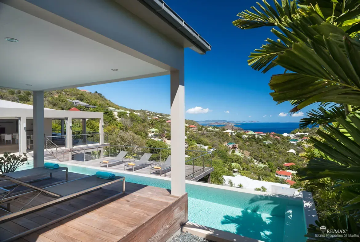 An infinity pool wraps around the weathered wooden terrace while turquoise towels and sun loungers set the tone for long afternoons in the sun. Below, lush green hills dotted with villas tumble toward the deep blue Caribbean and distant islands on the horizon.