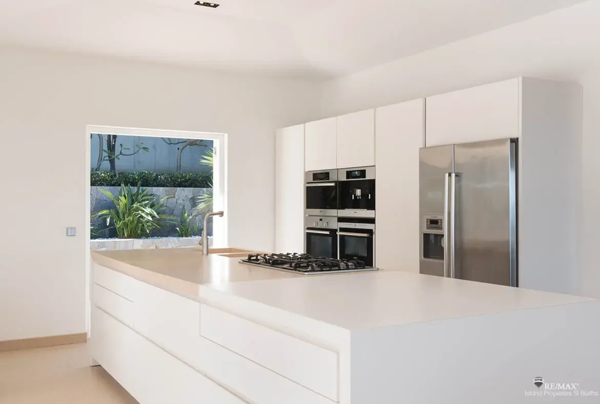 A sleek white island with integrated sink and cooktop sits opposite a wall of built-in ovens and a full-height refrigerator in this contemporary kitchen. Clean lines, pale flooring, and the open doorway to tropical greenery keep the whole space feeling fresh and effortless.