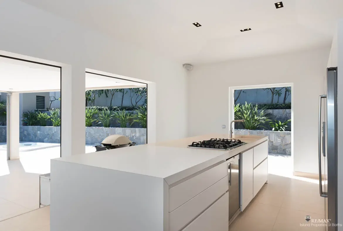 A sculptural white kitchen island with integrated cooktop, sink, and wine cooler anchors this open-air culinary space. Framed by tropical greenery and stone walls outside, it feels less like a kitchen and more like a sleek pavilion for long, easy vacation meals.