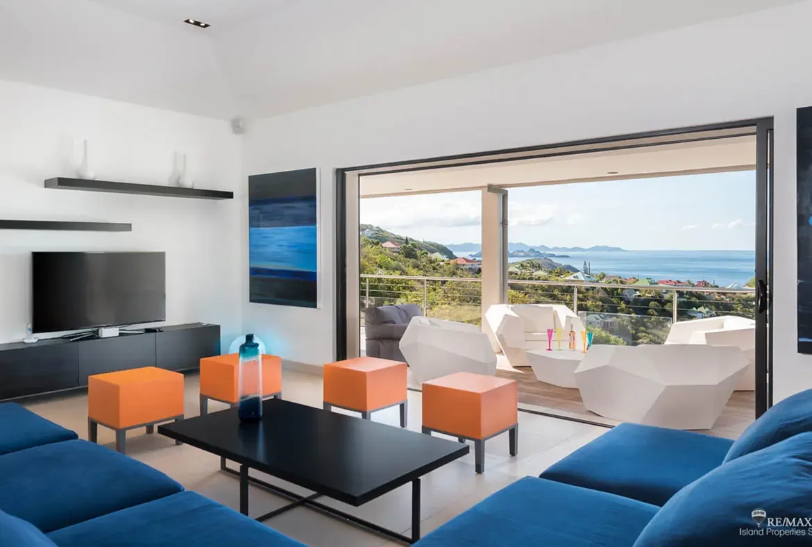 Two rich blue sectionals and bright orange ottomans bring bold energy to this contemporary living room, all pointed toward the terrace and the sea beyond. With hillside views dropping into the blue Caribbean, the whole space feels open, elevated, and designed for sunset lounging.