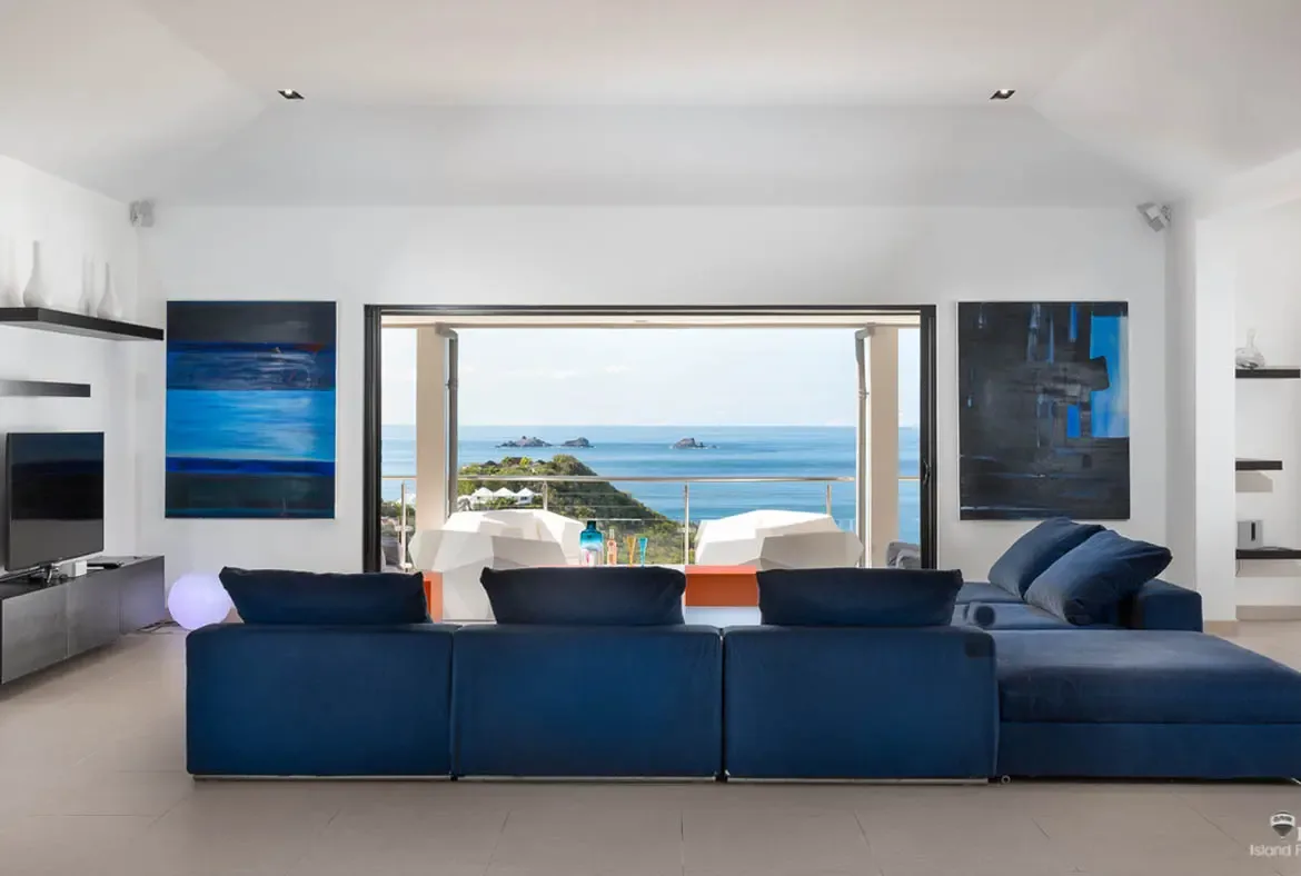 A navy modular sofa and cool blue artwork keep the focus on the real centerpiece here: the wide architectural opening that frames the Caribbean like a painting. White terrace chairs and an orange table add a crisp modern contrast to the sea and islands beyond.