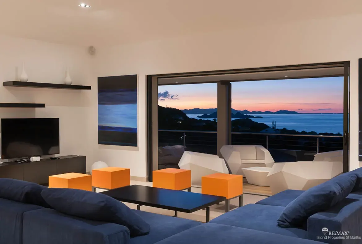 A deep blue sectional anchors this sleek living room, while the open terrace frames a dramatic Caribbean sunset over the sea and silhouetted hills. White sculptural chairs outside and warm orange accents inside make the whole scene feel effortless and cinematic.