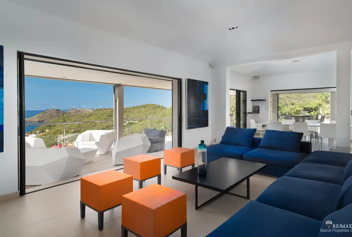Blue sectionals and bright orange cube stools create a playful, modern living space that opens straight onto the terrace. White lounge chairs and a bean bag face the hillside and ocean beyond, making the whole room feel like an extension of the view.