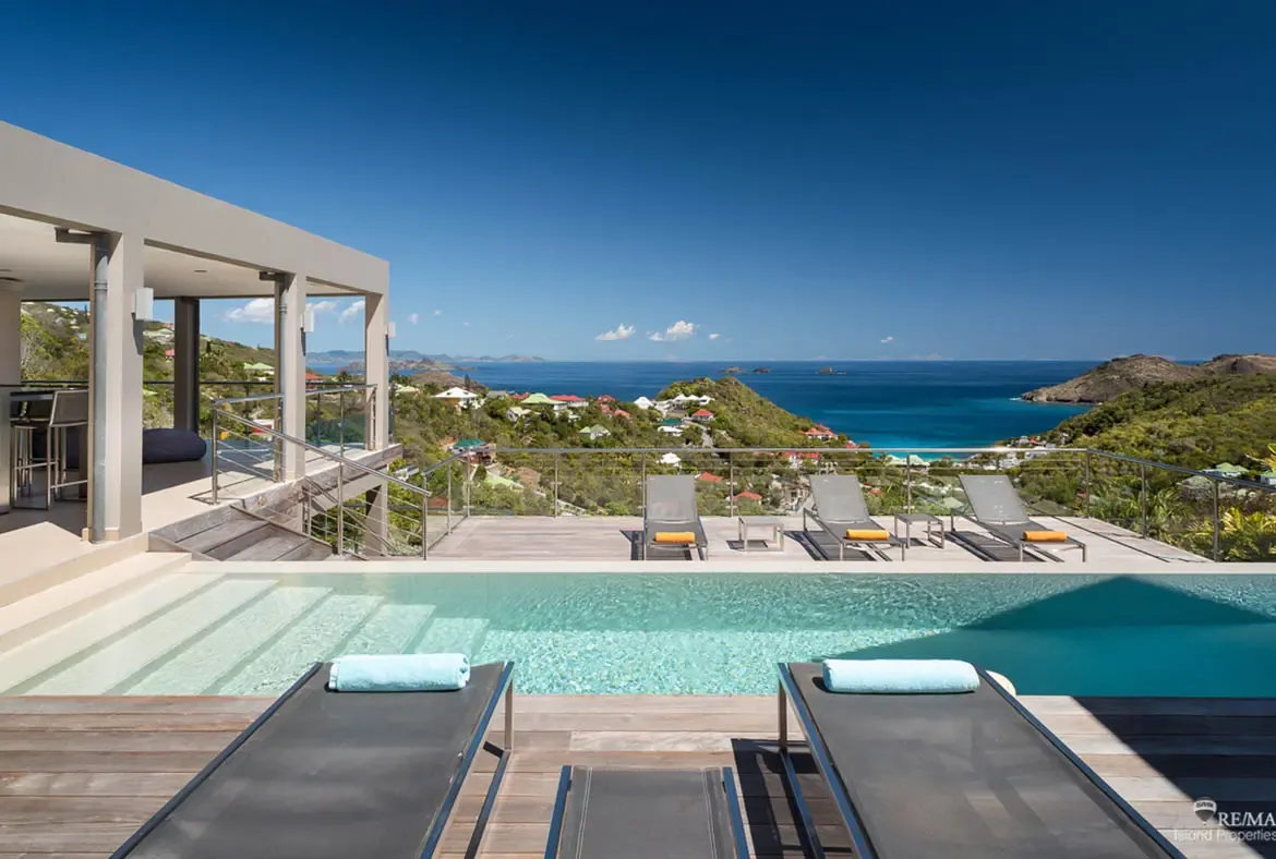 A long turquoise pool stretches across the wooden deck, framed by loungers with pale blue towels and glass railings that keep the view wide open. Beyond the hillside dotted with colorful homes, the ocean glows under a cloud-dusted Caribbean sky.