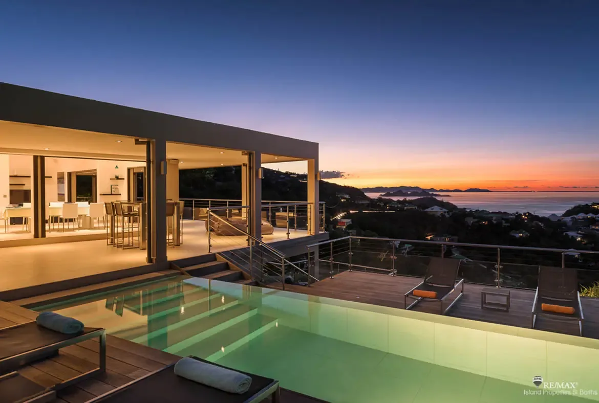 At twilight, the pool glows against the wooden deck as the villa opens wide to a softly lit modern interior. Beyond the terrace, green hills fall away to the sea while the sky turns gold, pink, and deep blue, delivering a classic St. Barths sunset moment.