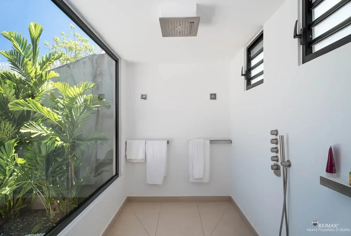 A ceiling-mounted rain shower and crisp white walls keep the design clean and spa-like, while the floor-to-ceiling window opens the whole room to tropical greenery outside. It’s a minimalist shower with a full island atmosphere.