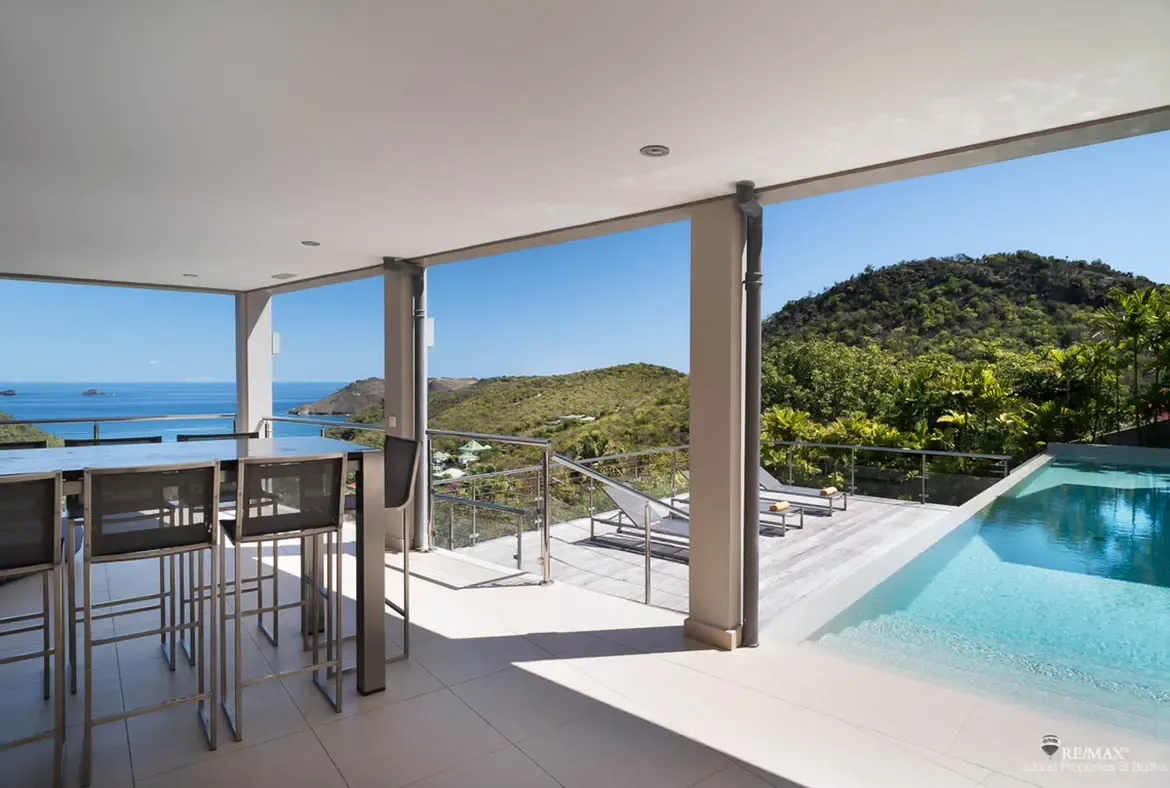 A covered terrace with sleek bar seating flows into a sun deck, infinity pool, and wide-open Caribbean views. With yellow-towel loungers facing the hills and sea, this setup feels made for lazy mornings and sunset aperitifs.