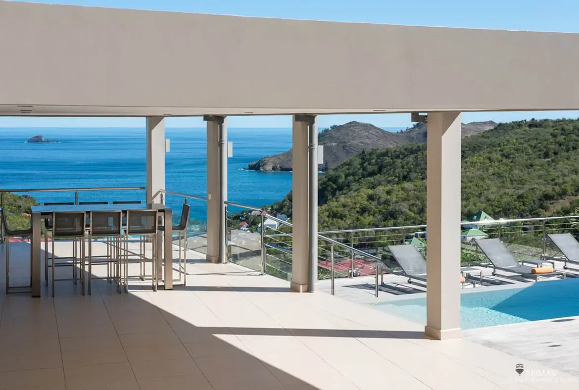 A shaded terrace dining area opens directly onto the infinity pool deck, where grey loungers with orange accents face the sea. With rolling green hills on one side and islands scattered across the horizon, the view stays dramatic from every angle.