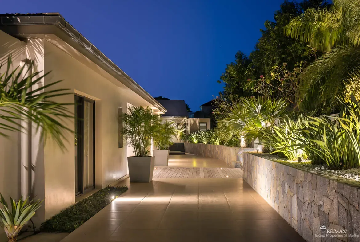 At dusk, warm lighting transforms this stone-lined walkway into a dramatic entrance framed by palms and sculpted tropical foliage. The mix of smooth tile, wood decking, and uplighted greenery gives the arrival a polished, cinematic feel.