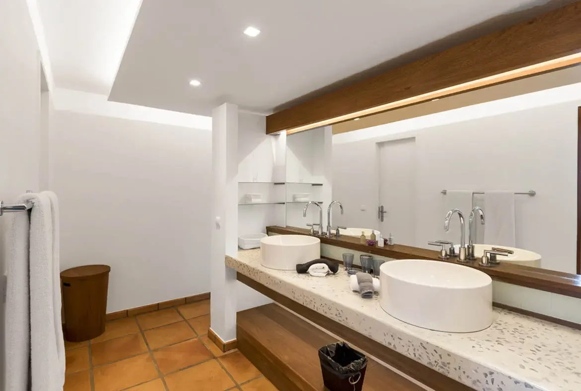 Two vessel sinks sit on a speckled stone counter under a wide dark-wood mirror with integrated lighting, while terracotta tiles and dark wood shelving keep this bathroom warm and grounded.