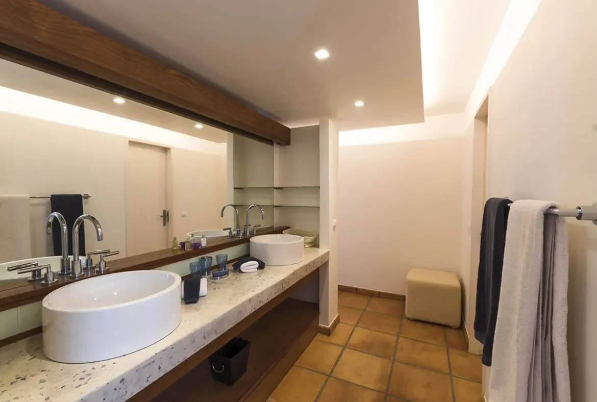 A sleek double vanity with vessel sinks sits below a dark-wood-framed mirror, while warm cove lighting and recessed spotlights keep this modern bathroom bright and polished.