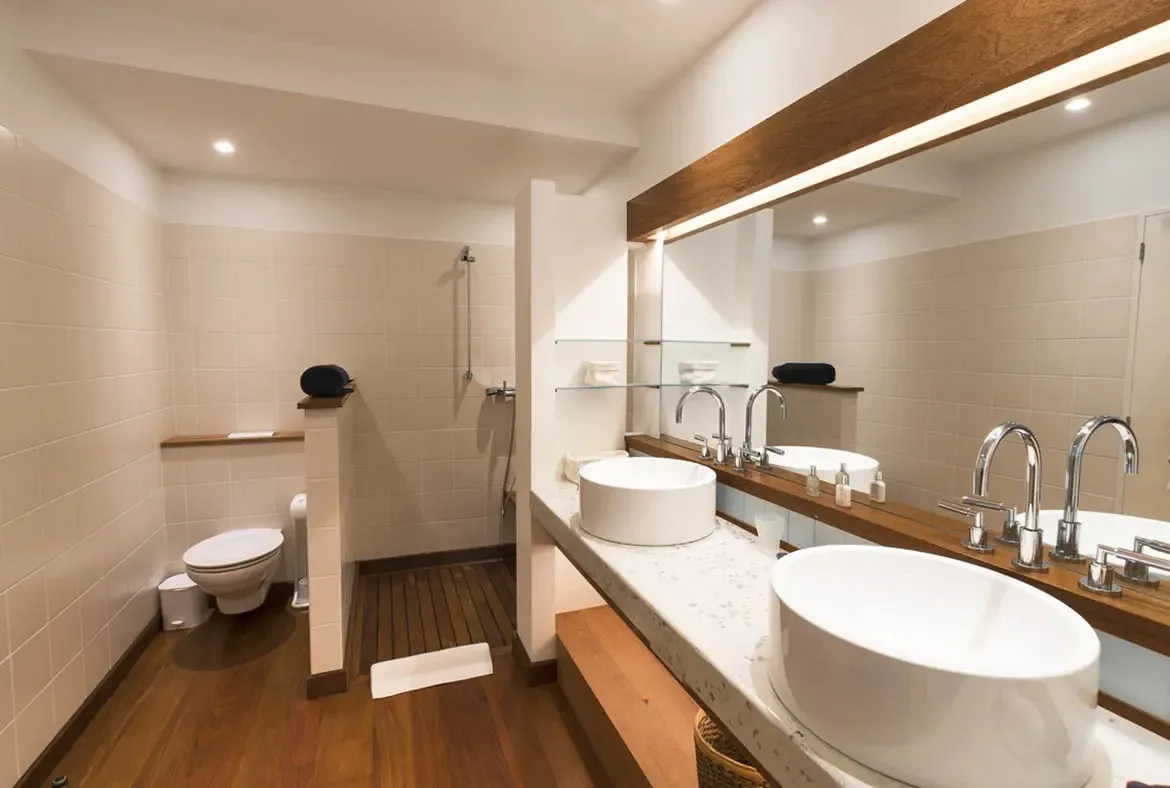 Two vessel sinks on a speckled stone counter sit below a wide dark-wood mirror, while warm wood floors and a walk-in shower with wood-slat flooring make this bathroom feel like a private spa.