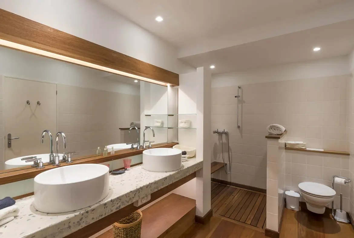Two vessel sinks under a warm wood-beam mirror pair with an open walk-in shower featuring a wood-slat floor and built-in bench, creating a bathroom that feels like a luxury spa retreat.