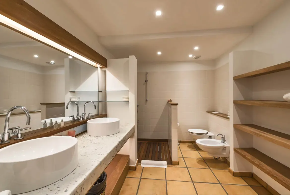 Two vessel sinks sit below a wide dark-wood-framed mirror with integrated lighting, while open wooden shelves and a walk-in shower with wood-slat flooring complete this polished bathroom.