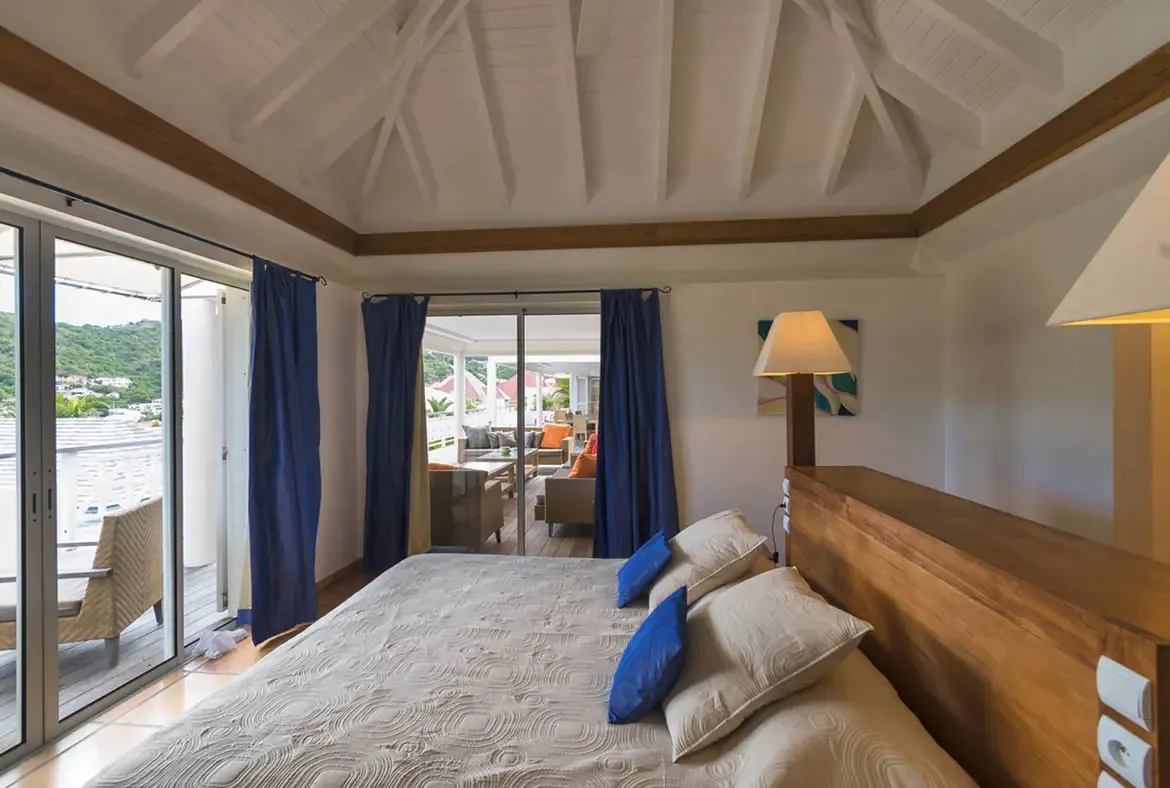Royal blue pillows and a custom wood headboard bring warmth to this airy bedroom, where glass doors open to a covered terrace with outdoor seating and a lush hillside panorama.
