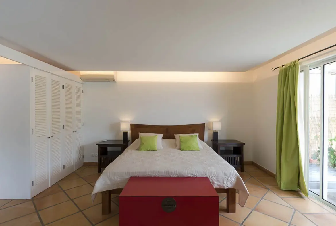 Lime-green pillows match floor-length curtains in this warm bedroom, where a red wooden chest adds Caribbean flair and glass doors lead out to a sunlit garden.