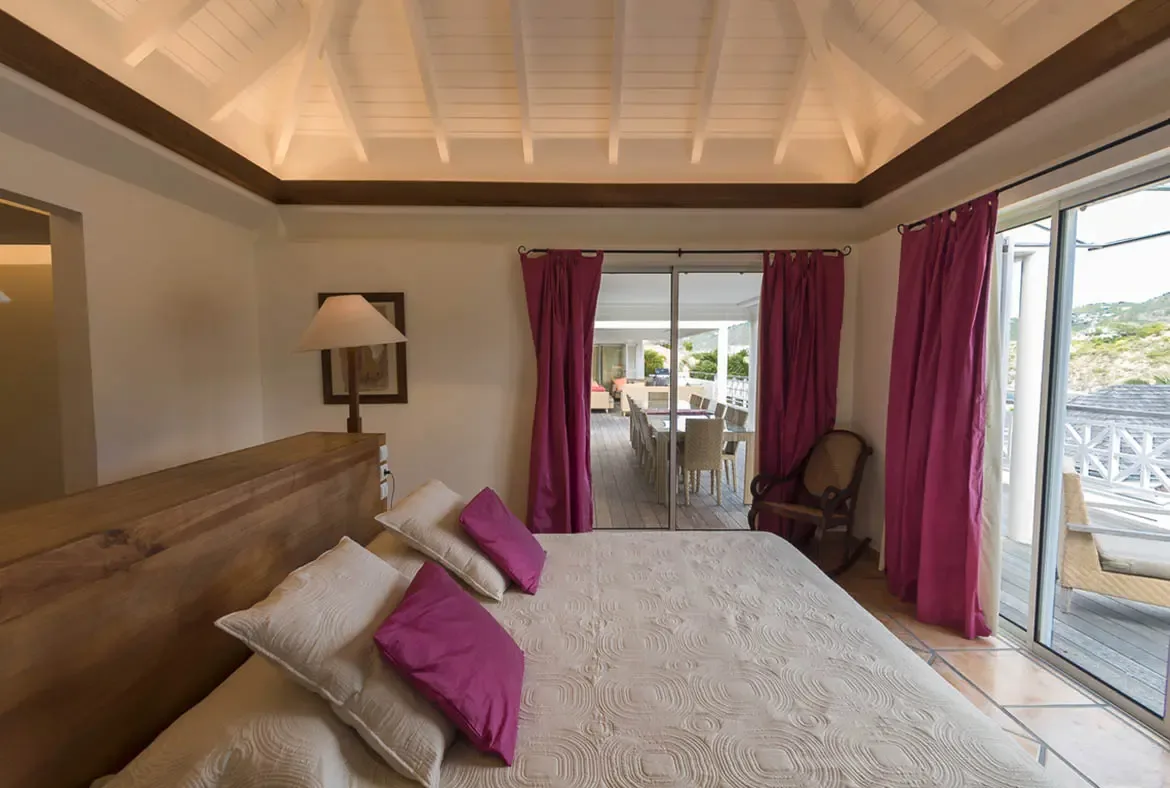 Magenta-purple pillows and curtains add personality to this serene bedroom with a rustic wood headboard, where glass doors open to an outdoor dining terrace and hillside views.