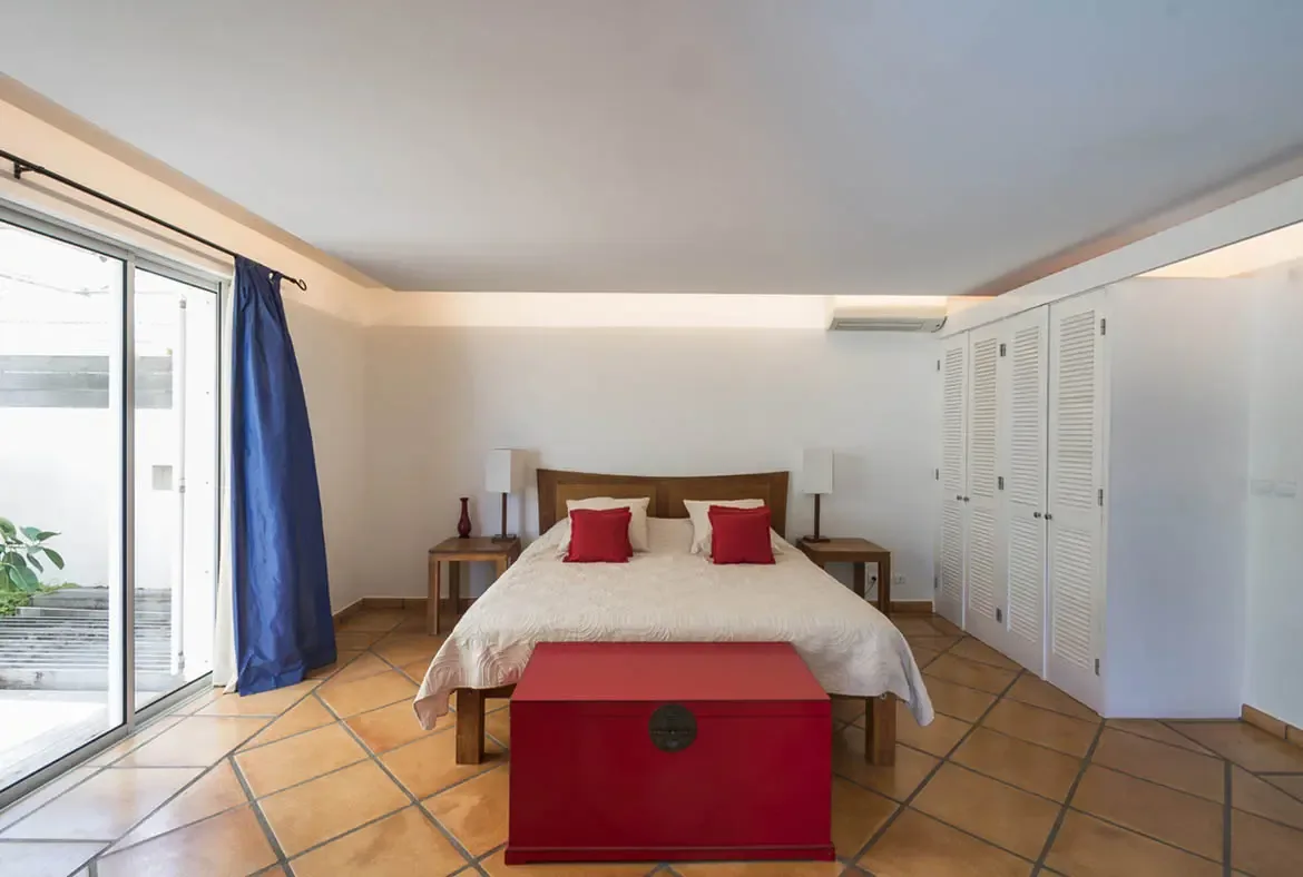 A bold red wooden chest at the foot of the bed and cobalt-blue curtains give this minimalist bedroom a vibrant Caribbean character, with glass doors opening to a leafy patio.