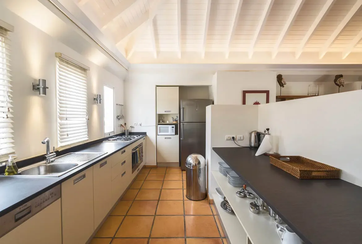A bright galley kitchen with terracotta tiles, black countertops, and a four-burner gas cooktop blends Mediterranean warmth with modern functionality beneath vaulted white rafters.