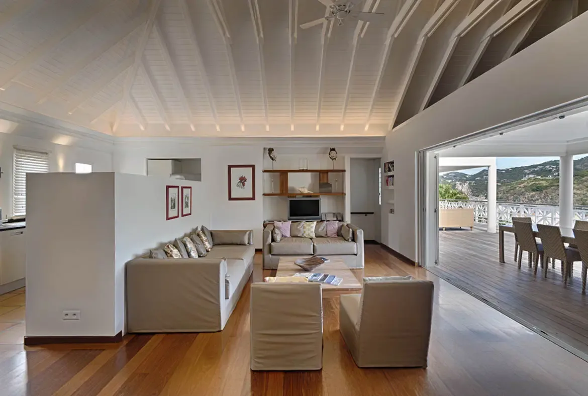 Folded-back glass doors erase the line between this vaulted living room and the wooden terrace, where an outdoor dining set faces green hills under a bright sky.