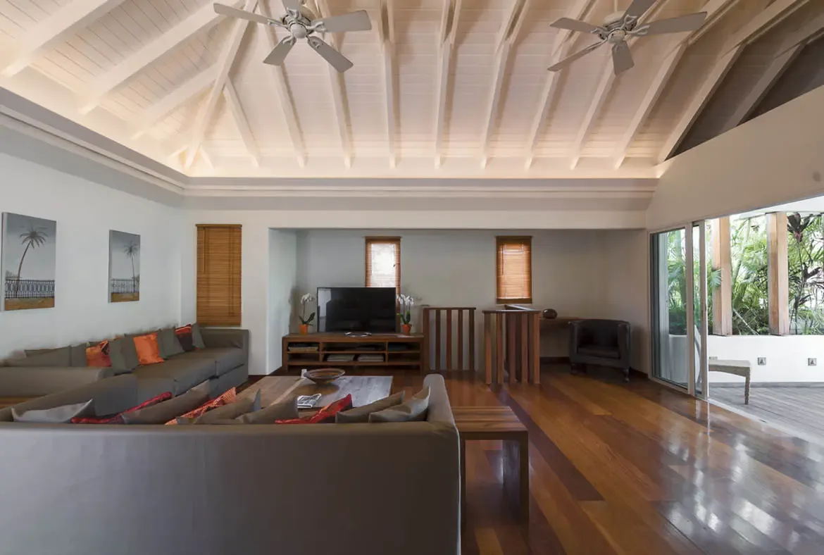 An L-shaped taupe sectional with orange and red pillows anchors this vaulted living room with polished hardwood floors, where open doors lead to a tropical deck under a bright Caribbean sky.
