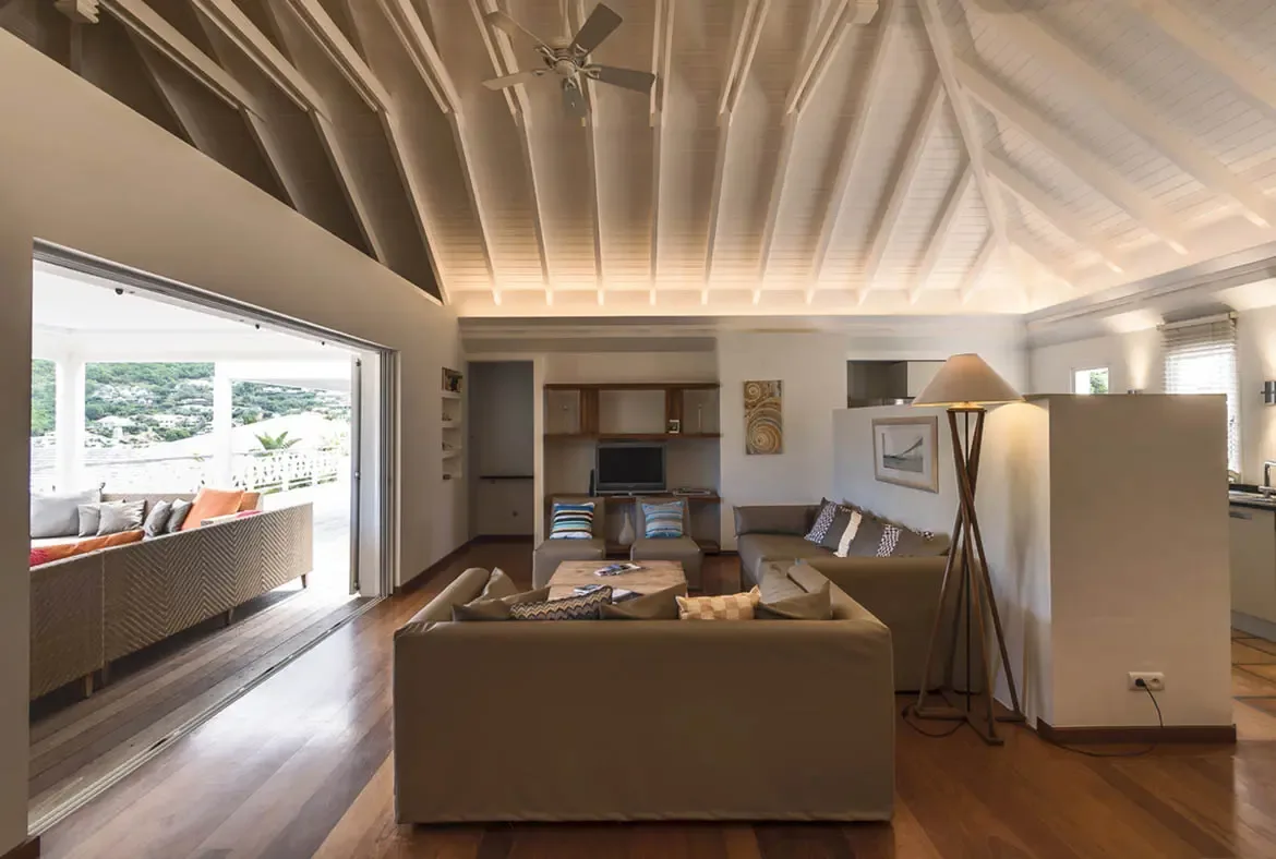A neutral taupe sectional and a warm hardwood floor anchor this vaulted-ceiling living room, where accordion glass doors open wide to a terrace with orange cushions and rolling green hills beyond.