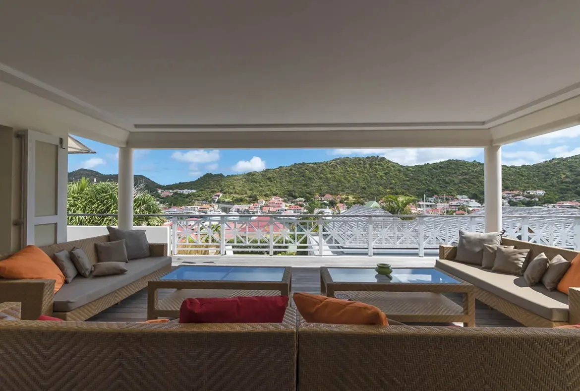Plush grey and orange cushions invite long afternoons on this covered terrace, where a white starburst railing frames a coastal town nestled around a harbor below green hills.