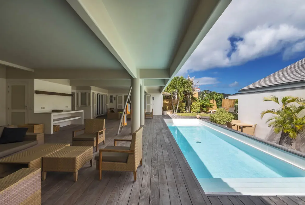 A long turquoise lap pool stretches across a weathered grey deck between two wings of the villa, with a shaded lounge area and potted palms creating a private hillside retreat.