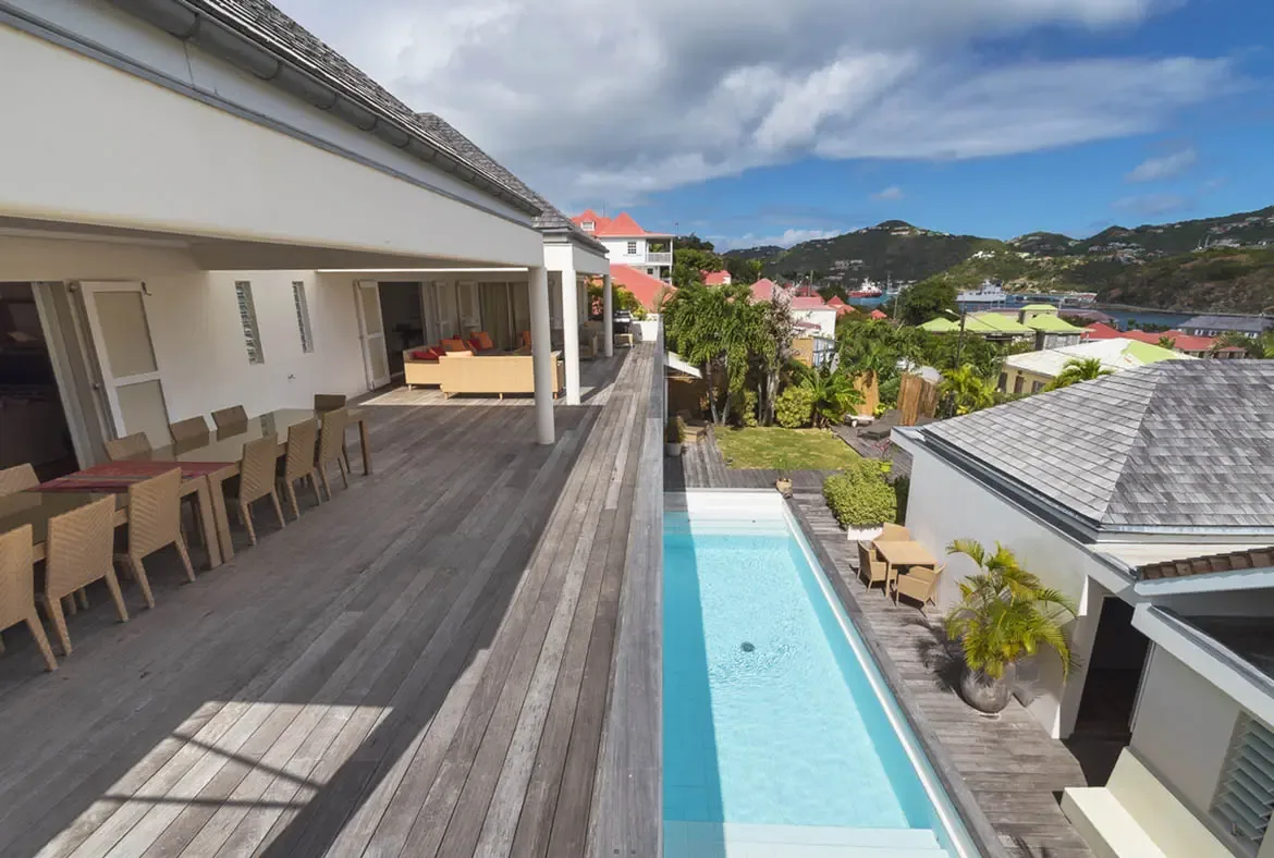 From above, a turquoise lap pool sits between two wings of the villa, with a dining terrace and lounge above, all overlooking a harbor town and green hills rising beyond.