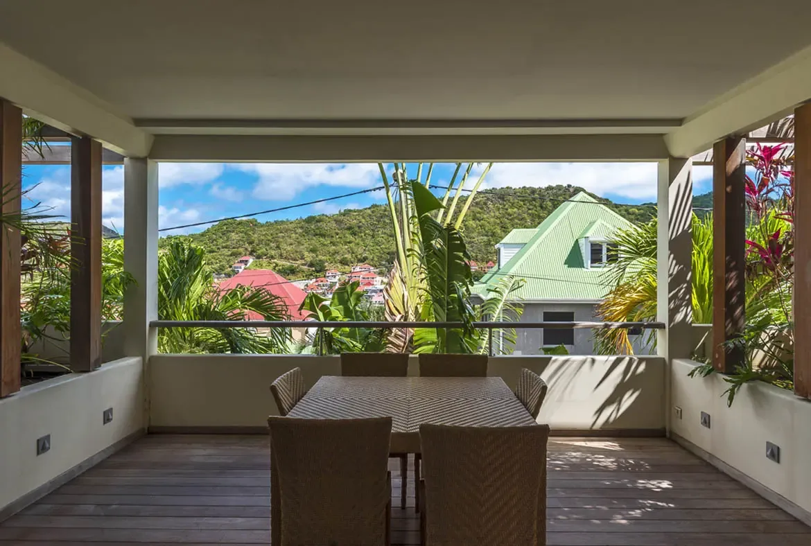 A woven dining table for six sits on a covered terrace with recessed lighting, where banana leaves frame a sweeping view of green hills dotted with colorful rooftops.