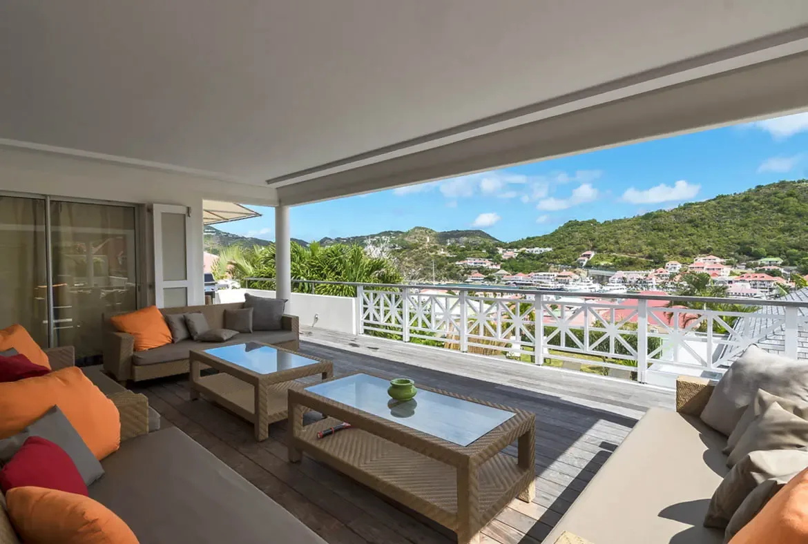 Deep wicker sectionals with burnt-orange cushions face a starburst railing and a sweeping harbor panorama, where green hills roll down to a bay scattered with anchored boats.