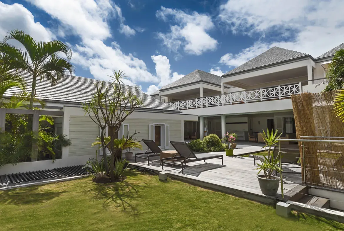 The villa's grey-shingled roofs and white starburst balconies rise above a manicured lawn with palms and tropical landscaping, framing a classic hillside St. Barths estate under a bright sky.