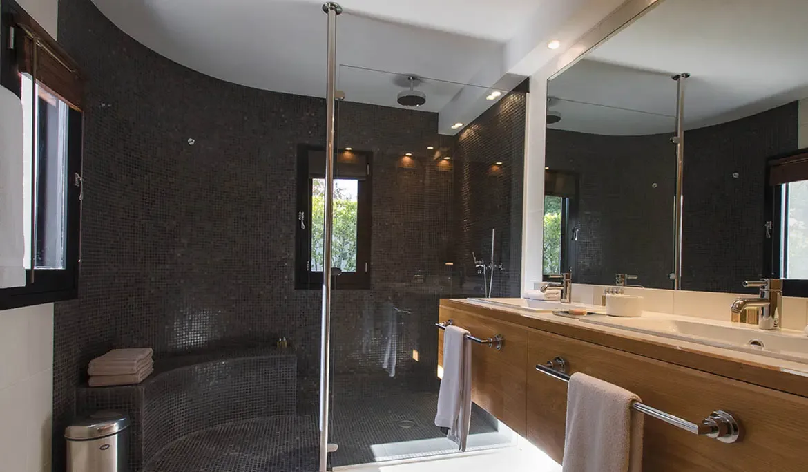 Dark mosaic tiles curve around a sunken shower with a built-in bench, while gold faucets top a warm wood vanity reflected in a wall-to-wall mirror.